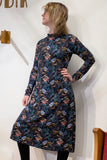 A person models the BUTIK KUBIK Kubik dress cinema birds maxi, a 60's inspired jersey dress with a colorful floral and bird pattern, posing in front of a white wall with one hand on their hip and looking to the side.