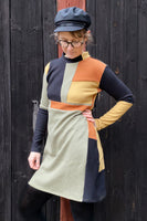 A person poses against a dark wooden background wearing glasses and a black cap. They proudly model the Kubik dress cinema patch unique M by BUTIK KUBIK, featuring 60s inspired green, orange, and black color-block sections crafted from sustainable Oeko-Tex fabric.