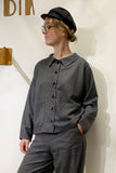 A person in glasses and a black cap wears the BUTIK KUBIK Kubik wool overshirt blouse houndstooth with matching pants, standing against a white wall, one hand in their pocket, looking to the side.