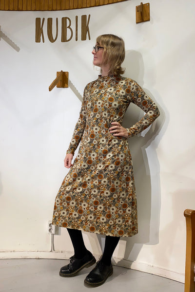 A person with glasses and shoulder-length hair wears the BUTIK KUBIK “Kubik dress cinema Gerbera maxi,” paired with black leggings and shoes, standing indoors near a wall sign that says “KUBIK.”.