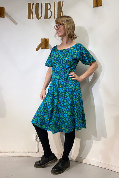 A woman in glasses models the Kubik dress Twinkle retro 1975 by BUTIK KUBIK, featuring a blue and green floral print, as she stands by a white wall with "KUBIK" written on it, smiling and posing with one hand on her hip.