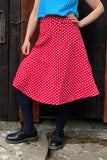 A person stands on a stone surface wearing a blue polka dot top, black tights, black shoes, and the BUTIK KUBIK Kubik skirt Twist Polka dot Red. A rustic wooden door is in the background.