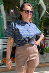A woman wearing the BUTIK KUBIK Kubik blouse Tetris green stands confidently outdoors with sunglasses and beige shorts, one hand on her hip. A mannequin and a red bicycle are visible in the background.