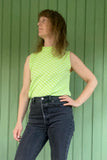 A sleeveless model stands before a green paneled wall, wearing the Kubik top cineast Limelight by BUTIK KUBIK—a light green, 60s-inspired knit with white polka dots—and high-waisted dark jeans, looking to the side with one hand on hip.
