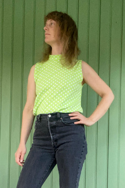 A sleeveless model stands before a green paneled wall, wearing the Kubik top cineast Limelight by BUTIK KUBIK—a light green, 60s-inspired knit with white polka dots—and high-waisted dark jeans, looking to the side with one hand on hip.
