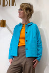 A person with short, wavy blond hair and glasses wears the BUTIK KUBIK Kubik Overshirt Strömma Lotta turkos over an orange shirt and brown checked pants, standing against a white wall and looking to the side.
