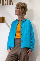 A person with short, wavy blond hair and glasses wears the BUTIK KUBIK Kubik Overshirt Strömma Lotta turkos over an orange shirt and brown checked pants, standing against a white wall and looking to the side.