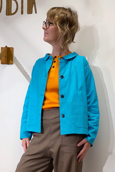 A person with short, wavy blond hair and glasses wears the BUTIK KUBIK Kubik Overshirt Strömma Lotta turkos over an orange shirt and brown checked pants, standing against a white wall and looking to the side.
