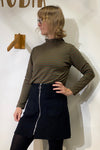 A blonde person with glasses poses sideways against a white wall with wooden decor, wearing the BUTIK KUBIK Kubik sweater stripes brown green and a black skirt featuring a front zipper and large pockets.