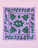 The Tropical Scandinavia scarf The Protector purple by BUTIK KUBIK hangs on a wall, displaying green "PROTECTOR OF FUN" text twice and graphics of turtles, a kettlebell, crescent moon, hand, and a large central eye.