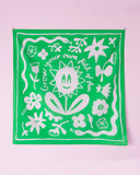 The BUTIK KUBIK Tropical Scandinavia scarf Grow your own, an organic cotton green scarf with playful white floral illustrations and the phrase "Grow your own field of fun," hangs against a light pink wall as a charming accessory.