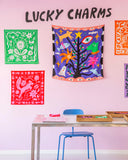 A display of vibrant patterned cloths, including the BUTIK KUBIK Tropical Scandinavia scarf Grow your own, hangs on a pink wall under "LUCKY CHARMS." A blue chair sits at a table with wooden trays holding these symbolic accessories.
