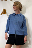 A woman with glasses wears the BUTIK KUBIK "Kubik blouse linen blue," an oversized button-up top made from 100% linen, paired with a black skirt featuring a front zipper. She stands against a white wall with a wooden hook behind her.