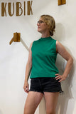 A person wearing the BUTIK KUBIK "Kubik top cineast green" and black shorts stands in front of a white wall with KUBIK written on it, resting one hand on their hip and looking to the side.