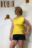 A woman stands in front of a white wall with KUBIK and wooden hooks, wearing the BUTIK KUBIK "Kubik top cineast yellow M"—a 60's inspired jersey A-line sleeveless top paired with black shorts. With one hand on her hip and looking to the side, she showcases this striking top, which is made in Sweden and blends retro charm with modern craftsmanship.