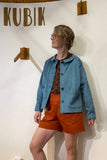 A person in glasses stands indoors wearing the BUTIK KUBIK Kubik corduroy overshirt in light petrol, a patterned shirt, and orange shorts. Wooden wall hooks and "KUBIK" appear on the white wall behind them.