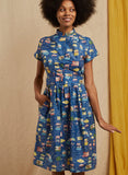 A woman with an afro smiles in the Palava Louise dress blue festival by BUTIK KUBIK, made from organic cotton satin with a fitted waist and pockets, standing against a neutral and mustard yellow fabric backdrop.