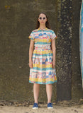 A woman poses against a textured wall in a Palava Louise dress multi beach local by BUTIK KUBIK. Her sunglasses catch the sun, complemented by blue sneakers. The sandy ground and a boat peeking from the right add an adventurous touch to this vibrant scene.