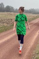 A woman in a BUTIK KUBIK Kubik dress cinema the leaf pairs it with navy leggings, tall white socks, and orange shoes as she walks along a dirt path in a grassy rural landscape, looking to her left.