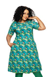 A woman with curly hair wears the Cissi och Selma Magda dress Kronblad Turkos by BUTIK KUBIK, featuring turquoise, green, and yellow patterns. She stands confidently with one hand in her pocket against a white background.