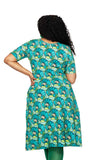 A woman with curly hair stands with her back to the camera, wearing the Cissi och Selma Magda dress Kronblad Turkos by BUTIK KUBIK, featuring a blue and green geometric floral pattern, paired with green tights and one hand on her hip.