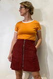 A person wearing glasses, an orange ringer T-shirt, and the BUTIK KUBIK “Kubik corduroy zip skirt nr 2,” a burgundy A-line skirt with a front zipper, stands against a plain light-colored wall, looking to their left.