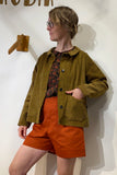A person with glasses and light hair pulled back wears the BUTIK KUBIK Kubik corduroy overshirt in Olive, paired with a patterned black and red shirt and orange shorts, standing against a white wall with wooden decor.