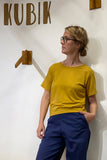 A woman with glasses, dressed in the BUTIK KUBIK Kubik box tee mustard modal and blue pants, stands with one hand in her pocket in front of a wall featuring wooden text that reads KUBIK.