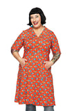 A smiling person with short black hair wears the Cissi och Selma dress Monica Midsommardans Röd by BUTIK KUBIK. They have tattoos, stand with hands in pockets, and pose against a white background.