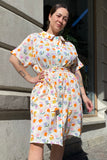 A woman with arm tattoos stands confidently outdoors against a gray stone wall, wearing the BUTIK KUBIK Kubik blouse dress floral muslin, a belted short-sleeve dress with a colorful flower pattern, in sunlight.