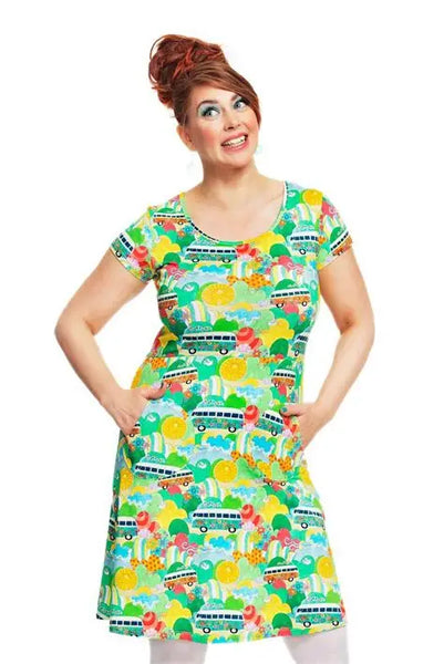 A smiling woman with red hair in an updo poses with hands in the pockets of the BUTIK KUBIK Cissi och Selma dress Asta Festival, featuring playful vans, rainbows, and sun prints on white organic cotton.