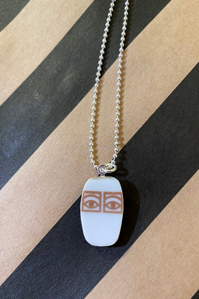 The Saved & remade necklace Porslin by BUTIK KUBIK features a silver-plated chain with a white rectangular pendant, showing two brown outlined eyes, crafted from recycled porcelain. Displayed on a black and tan diagonal striped background.