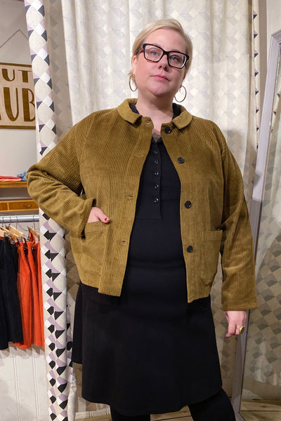 A person wearing glasses, a black dress, and the BUTIK KUBIK Kubik corduroy overshirt in olive stands indoors with one hand in a pocket. Patterned curtains, hanging clothes, and a sign appear in the background.
