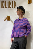 A person in a black cap, glasses, gray pants, and the Kubik cotton shirt striped by BUTIK KUBIK stands before a wall with KUBIK and wooden hooks, looking to the side.
