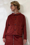 A person with short blond hair and glasses wears the BUTIK KUBIK Kubik corduroy overshirt in burgundy, featuring black buttons and large front pockets, styled with a matching skirt. They stand against a plain white wall, looking to the side.