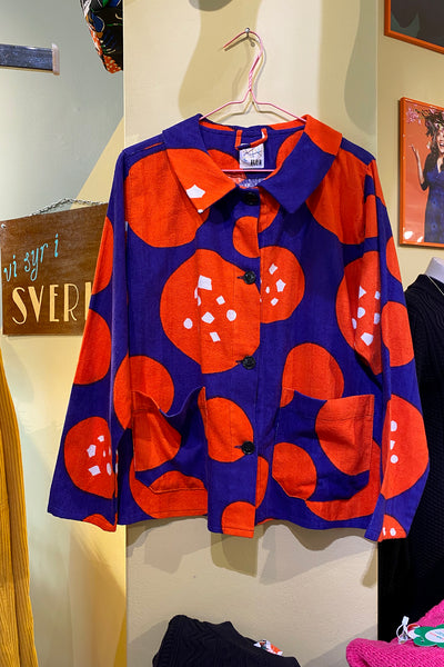 The BUTIK KUBIK Kubik Overshirt Vintage Terry features bold red and purple geometric patterns on deadstock fabric—large red circles with white atop deep purple—displayed on a pink hanger against a light yellow wall.