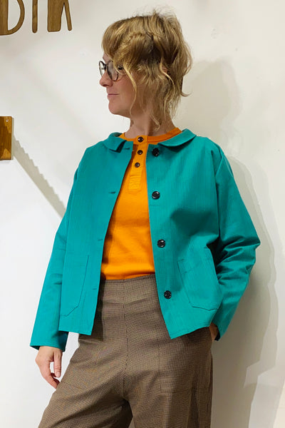 A person with short, wavy hair and glasses wears the BUTIK KUBIK Kubik Overshirt Strömma Lotta grön, crafted from vintage deadstock fabric, over an orange shirt and brown plaid pants against a plain white background.