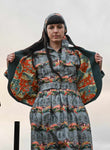 A woman with long dark hair wears the BUTIK KUBIK Palava Dress Cynthia leaping deer—a patterned dress—while holding open a matching coat to show its bright orange nature-patterned lining beneath an overcast sky.