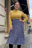 A woman smiles on a cobblestone street, wearing a yellow polka-dot top and the BUTIK KUBIK Kubik skirt Twist Pärlsocker, a navy blue 50s-inspired skirt with white dots. Cloudy day; buildings and sidewalk in the background.