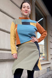 A woman stands outside smiling, hand on hip, wearing the BUTIK KUBIK Kubik cropped sweater waffle patch size L—60's inspired with bold geometric blue, yellow, orange, green, and black patterns—made in Sweden.
