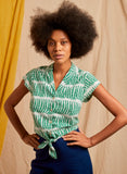 A woman with a natural afro, hands on hips, wears the Palava Peggy blouse green peas by BUTIK KUBIK—50s-inspired in a green and white organic cotton-linen blend—with navy pants, posed before a beige and yellow fabric backdrop.