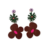 The Törmi Kimara earrings by BUTIK KUBIK feature lightweight lavender flowers, green palm-like leaves, and bold brown blooms with pink centers on white. Eco-friendly packaging included.