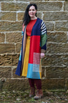 A woman with long dark hair and a neutral expression stands against a stone wall, wearing the Palava Polly Cardigan Coat by BUTIK KUBIK over a red top and yellow skirt, paired with burgundy tights and brown lace-up boots for an ethical fashion look.