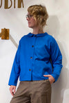 A person with wavy blonde hair and glasses wears the Kubik Overshirt Strömma Lotta blå by BUTIK KUBIK, a bright blue Swedish-made shirt with two front pockets, paired with brown plaid pants, standing against a white wall.