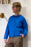 A person with wavy blonde hair and glasses wears the Kubik Overshirt Strömma Lotta blå by BUTIK KUBIK, a bright blue Swedish-made shirt with two front pockets, paired with brown plaid pants, standing against a white wall.