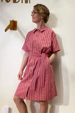 A woman in glasses wears the BUTIK KUBIK Kubik blouse dress in pink stripes, complete with a matching belt, and stands against a white wall, looking to the side with one hand in her pocket.