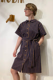 A woman wearing glasses stands against a white wall, dressed in the BUTIK KUBIK Kubik blouse dress brown/blue stripe with a matching waist belt and her hair pinned up.