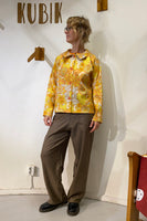 A person with short, wavy hair and glasses stands indoors wearing a yellow patterned Kubik Overshirt retro by BUTIK KUBIK, brown pants, and black shoes. They look to the side, with a sign reading KUBIK on the wall behind them.