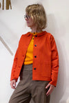 A person with wavy blonde hair and glasses wears the Kubik Overshirt Strömma Lotta röd by BUTIK KUBIK over a yellow shirt and brown checkered pants, standing against a plain white background.
