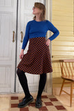 A woman poses confidently indoors on a checkered rug, wearing glasses, a blue long-sleeve top, black tights and shoes, and the BUTIK KUBIK Kubik skirt Twist short checkered—a 50s-inspired brown-and-black checkered skirt.